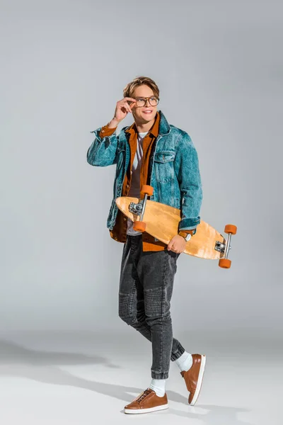 Joven feliz hombre en chaqueta de mezclilla posando con monopatín en gris - foto de stock