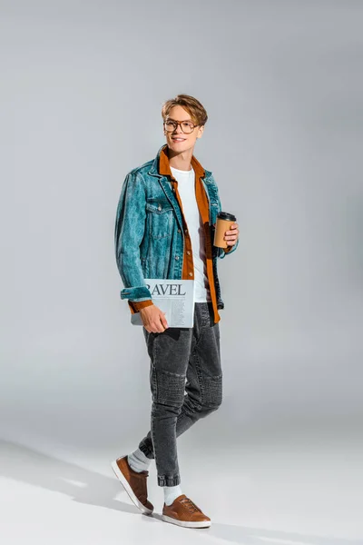 Guapo hipster en chaqueta de mezclilla sosteniendo café para ir y periódico de viaje en gris - foto de stock