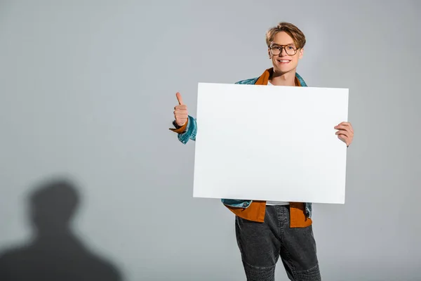 Handsome smiling hipster holding empty board and showing thumb up on grey — Stock Photo