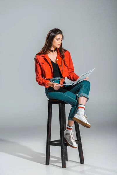 Mujer joven con estilo leyendo el periódico de viaje mientras está sentado en el taburete en gris - foto de stock