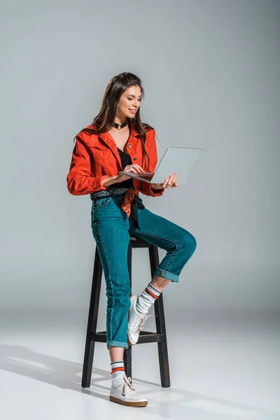 Приваблива дівчина використовує ноутбук і сидить на табуреті на сірому — стокове фото