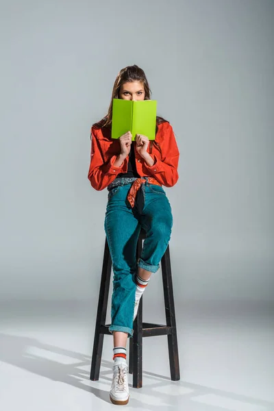 Atractivo estudiante elegante sosteniendo libro verde mientras está sentado en el taburete en gris - foto de stock