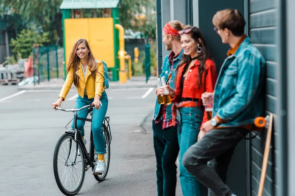Atractiva chica elegante montar en bicicleta cerca de amigos con botellas de cerveza en la calle - foto de stock