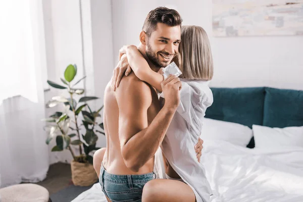 Sorridente giovane uomo che tiene preservativo mentre abbraccia la ragazza in camera da letto e guardando la fotocamera — Foto stock