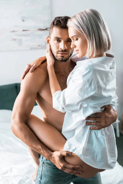 Hemdloser schöner Mann trägt seine Freundin im Schlafzimmer und schaut in die Kamera — Stockfoto