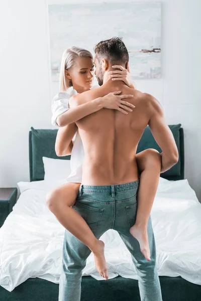 Вид ззаду без сорочки м'язистий чоловік, що носить свою дівчину в спальні — стокове фото