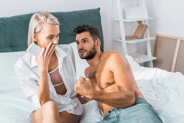 Chocado jovem mulher segurando smartphone de seu namorado confuso enquanto ele deitado na cama perto dela — Fotografia de Stock