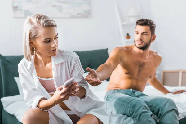 Jealousy young woman using smartphone of her confused boyfriend while he sitting on background in bedroom — Stock Photo