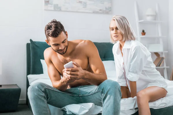 Celos joven usando el teléfono inteligente de su novia mientras ella está sentada en el fondo en el dormitorio - foto de stock