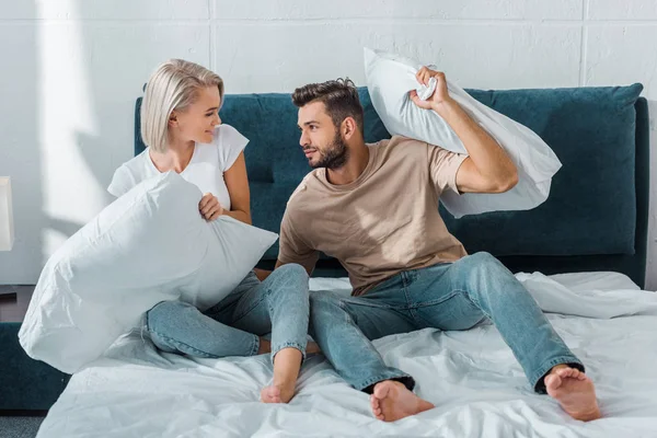 Cheerful boyfriend and girlfriend fighting with pillows on bed in bedroom — Stock Photo