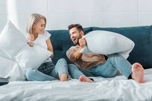Счастливая молодая пара борется с подушками на кровати в спальне — стоковое фото