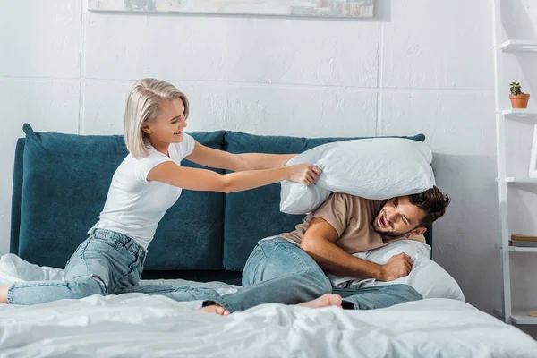 Счастливая девушка избивает парня подушкой на кровати в спальне — стоковое фото