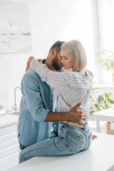 Afetuoso jovem casal abraçando na cozinha, namorada sentada no balcão da cozinha — Fotografia de Stock