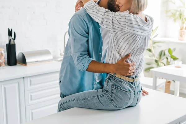 Imagem cortada de jovem casal abraçando na cozinha, namorada sentada no balcão da cozinha — Fotografia de Stock