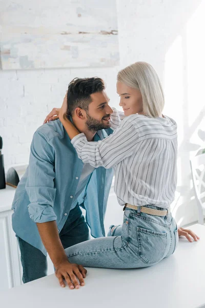 Attractive girlfriend sitting on kitchen counter and hugging handsome boyfriend in kitchen — Stock Photo