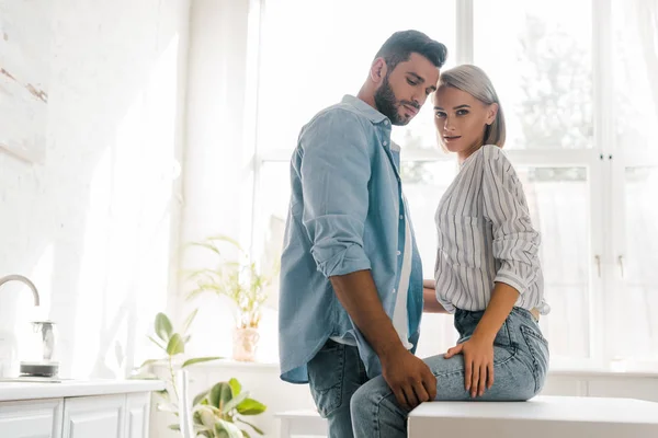 Side view of passionate young couple hugging in kitchen, girlfriend looking at camera — Stock Photo