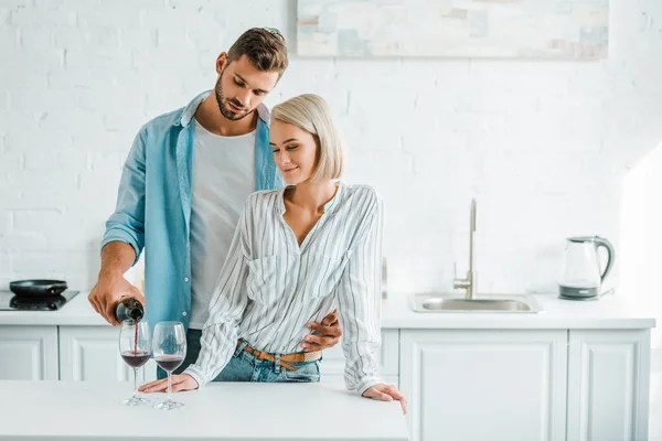 Bello fidanzato abbracciare fidanzata da indietro in cucina e versando vino da bottiglia — Foto stock