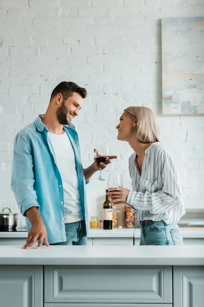 Souriant jeune couple parlant, tenant des verres de vin rouge et se regardant dans la cuisine — Photo de stock