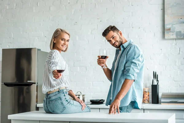 Sonriente joven pareja mostrando vasos de vino tinto y mirando a la cámara en la cocina - foto de stock