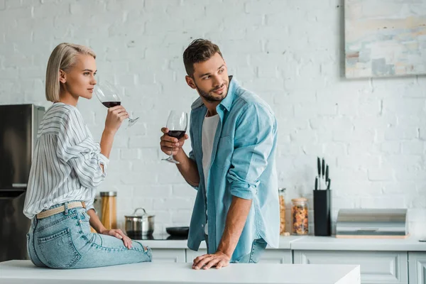 Pareja joven bebiendo vino tinto y mirando hacia otro lado en la cocina - foto de stock