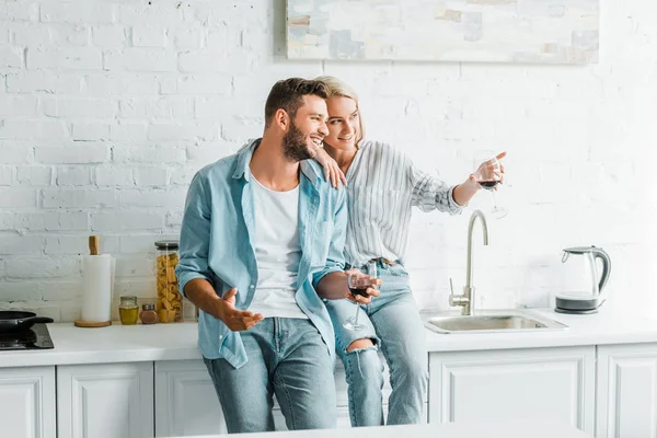 Sonriente novia sosteniendo vaso de vino y señalando algo a su novio en la cocina - foto de stock