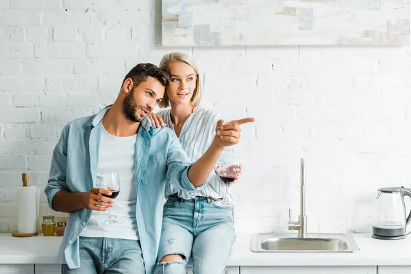 Boyfriend holding glass of wine and pointing on something to girlfriend in kitchen — Stock Photo