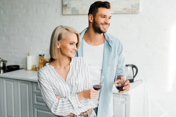 Sonriente joven pareja sosteniendo vasos de vino tinto y mirando hacia otro lado en la cocina - foto de stock