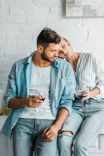 Novia sosteniendo vaso de vino y apoyándose en guapo novio en cocina - foto de stock