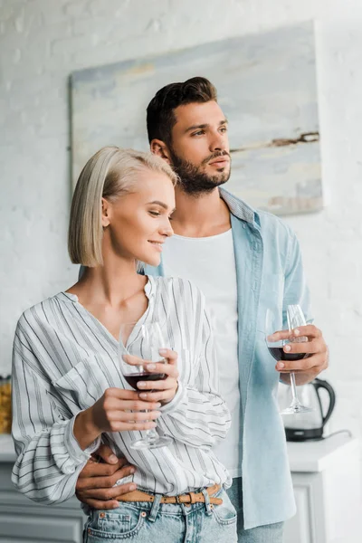 Jovem casal abraçando, segurando copos de vinho tinto e olhando para longe na cozinha — Fotografia de Stock