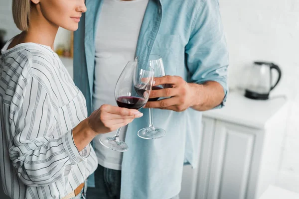 Imagen recortada de pareja joven tintineo con gafas de vino en la cocina - foto de stock