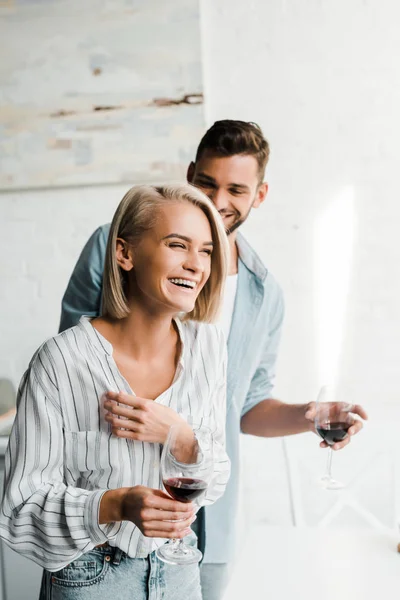 Jovem casal segurando copos de vinho e rindo na cozinha — Fotografia de Stock