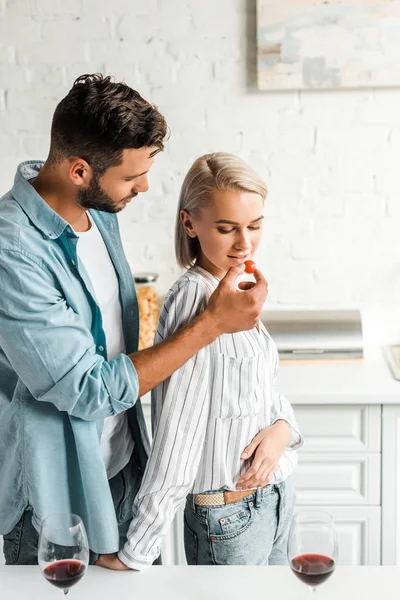 Handsome boyfriend feeding girlfriend with cherry tomato in kitchen — Stock Photo