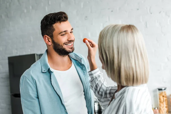 Girlfriend feeding smiling handsome boyfriend with cherry tomato in kitchen — Stock Photo