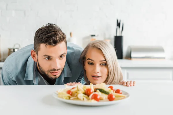 Surprised girlfriend and boyfriend looking out from table and looking at plate with salad in kitchen — Stock Photo