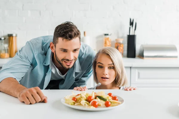 Jovem casal surpreso olhando para fora da mesa e olhando para prato com salada na cozinha — Fotografia de Stock
