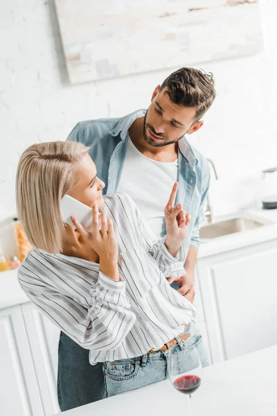 Jovem namorada falando por smartphone e mostrando um dedo até namorado na cozinha — Fotografia de Stock