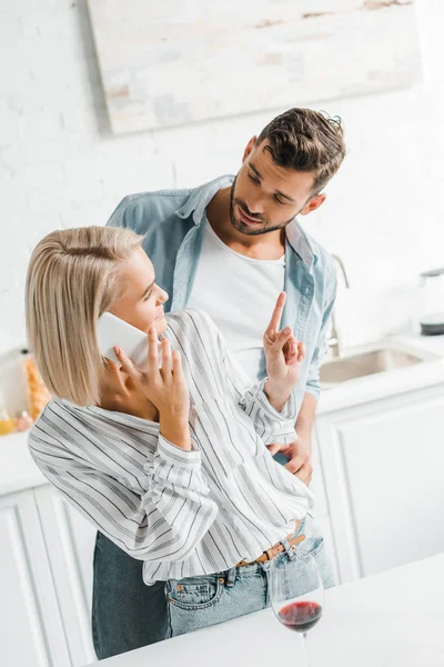 Joven novia hablando por teléfono inteligente y mostrando un dedo hasta novio en la cocina - foto de stock