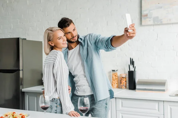 Веселая молодая пара делает селфи со смартфоном на кухне — стоковое фото