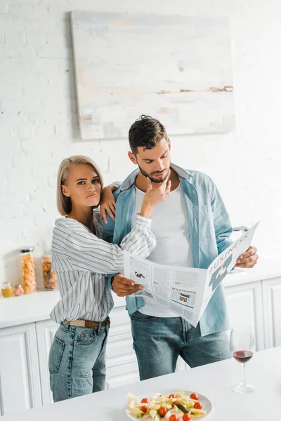 Boyfriend reading newspaper and grimacing girlfriend touching his chin in kitchen — Stock Photo