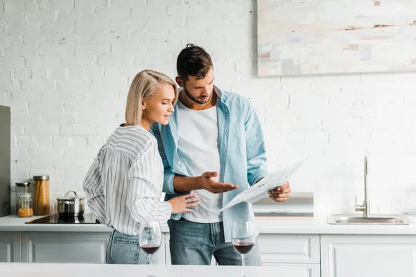 Handsome boyfriend pointing on newspaper to smiling blonde girlfriend in kitchen — Stock Photo