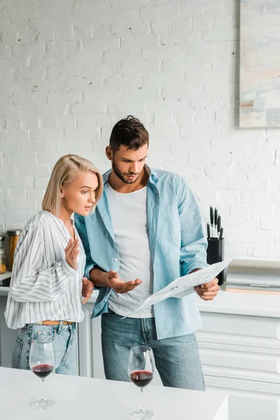 Guapo novio apuntando en periódico a sorprendido novia en cocina - foto de stock