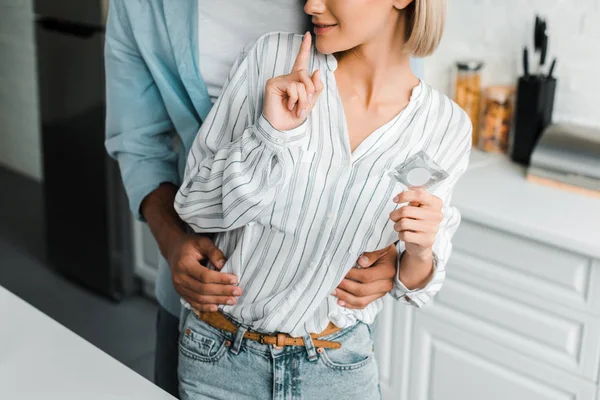 Cropped image of girlfriend showing silence gesture and holding condom in kitchen — Stock Photo