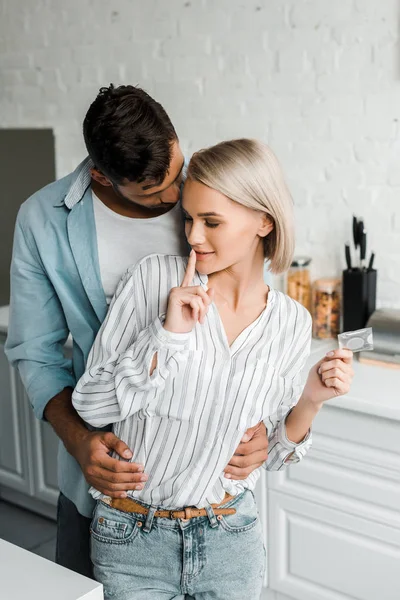 Attractive girlfriend showing silence gesture and holding condom in kitchen — Stock Photo