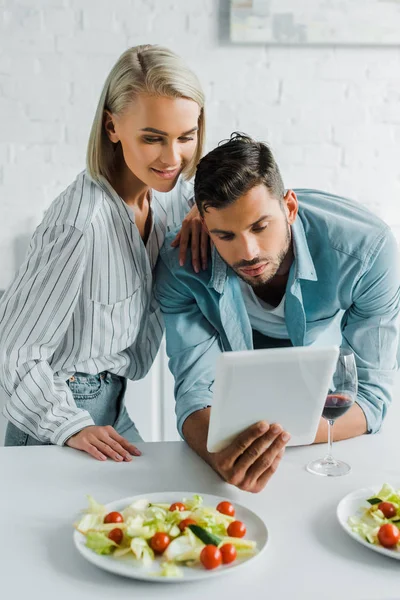 Young couple using tablet in kitchen, salad on tabletop — Stock Photo