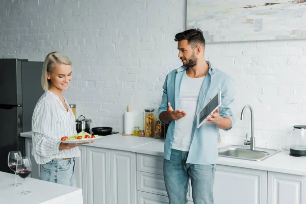 Novio señalando en la tableta a la novia sonriente con plato en la cocina - foto de stock