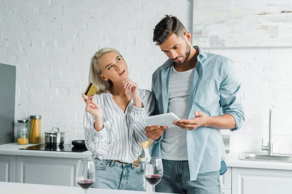 Novio sosteniendo tableta y novia pensativa de pie con tarjeta de crédito en la cocina - foto de stock