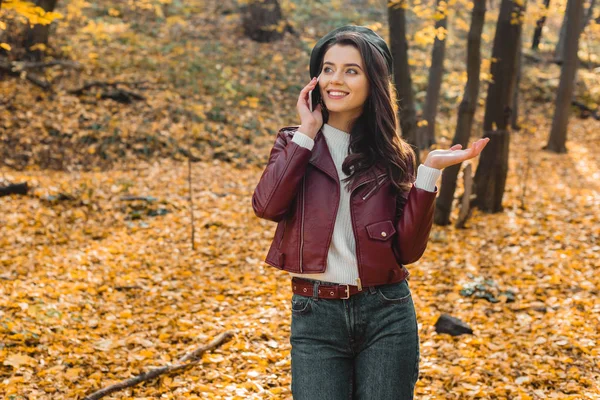 Jovem mulher elegante em jaqueta de couro falando no smartphone e gesticulando à mão no parque outonal — Fotografia de Stock
