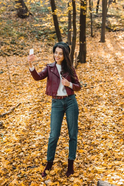 Attractive girl in stylish leather jacket and beret taking selfie on smartphone in yellow forest — Stock Photo