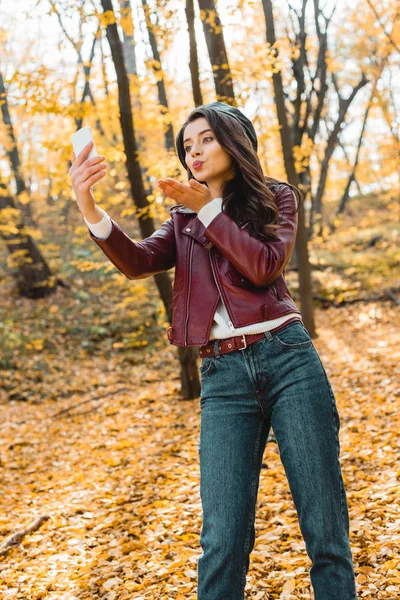 Low angle view of beautiful woman in stylish leather jacket blowing air kiss and taking selfie on smartphone in forest — Stock Photo