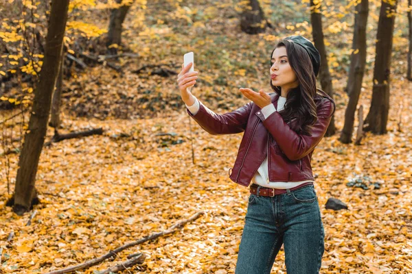 Jeune femme dans la veste en cuir élégant souffler baiser d'air et de prendre selfie sur smartphone dans le parc automnal — Photo de stock